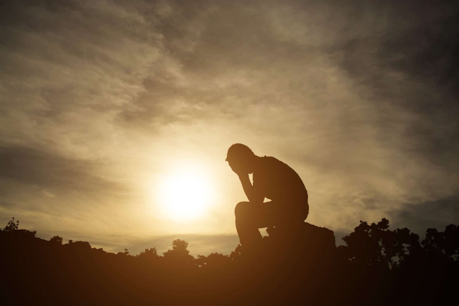 Silhouette of a person sitting in a pensive, hunched pose against a bright sunset to represent overcoming addiction.