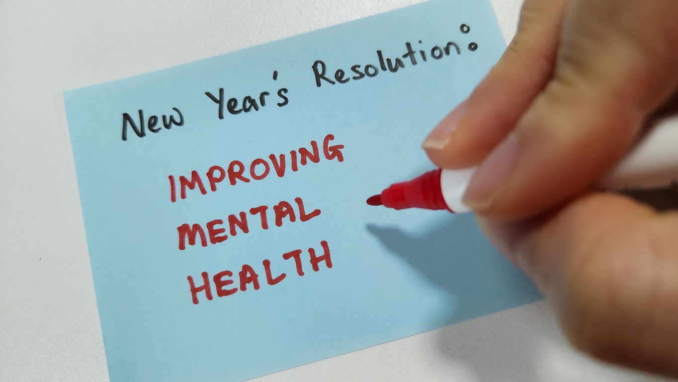Hand with red marker by a light blue sticky note reading “New Year’s Resolution: IMPROVING MENTAL HEALTH” on white.