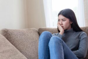 Young woman sitting on a sofa with her hand on her chin, looking away with a worried or depressed expression.