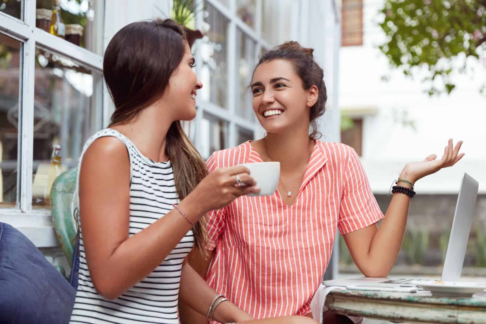 Two women sit outside at a cafe next to a laptop.