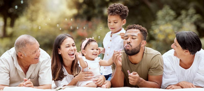 A family lays across a blanket outside, smiling and blowing bubbles.