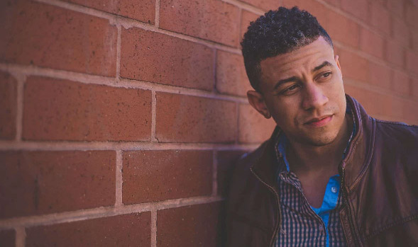 A young man, looking off into the distance, leans against a brick wall.