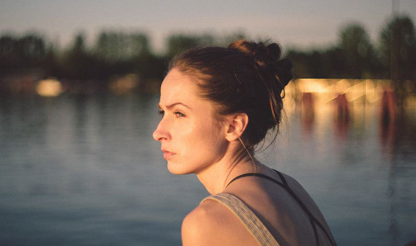 A young woman stands by a pond in the background. She looks to the left contemplatively staring into the distance.