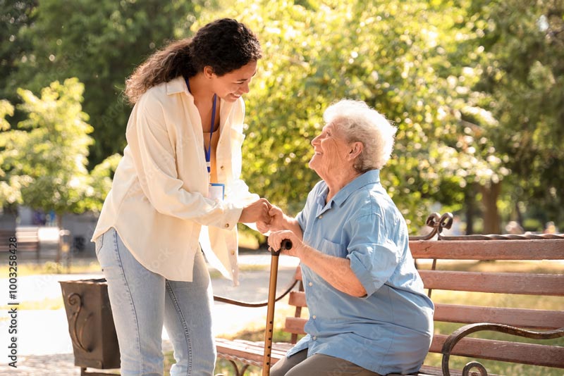 Two happy women, one younger and one older, converse near a park bench on a sunny day.