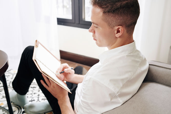 A young man seated on the floor leans against a couch. He holds open a journal and holds a pen in his right hand.