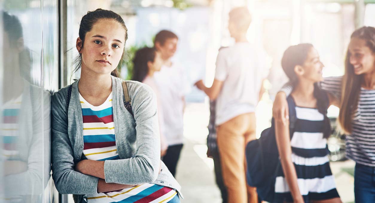 A teen, school-age girl leans against a wall at school. She appears sad. Other students in the background appear happy and smiling.