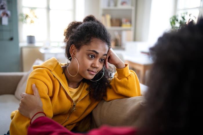 A young woman rests an elbow on the back of the couch. She stares into the distance. A loved one touches her arm to comfort her.